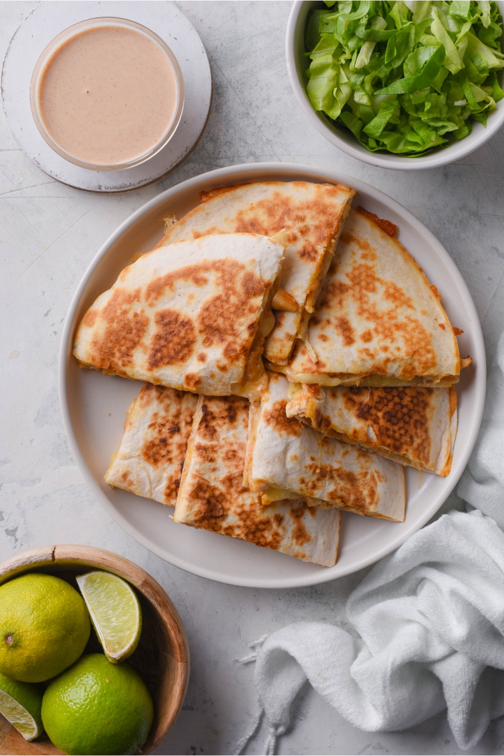 A plate of chicken quesadilla wedges piled on top of each other next to a bowl of quesadilla sauce, a bowl of limes, and a bowl of lettuce.