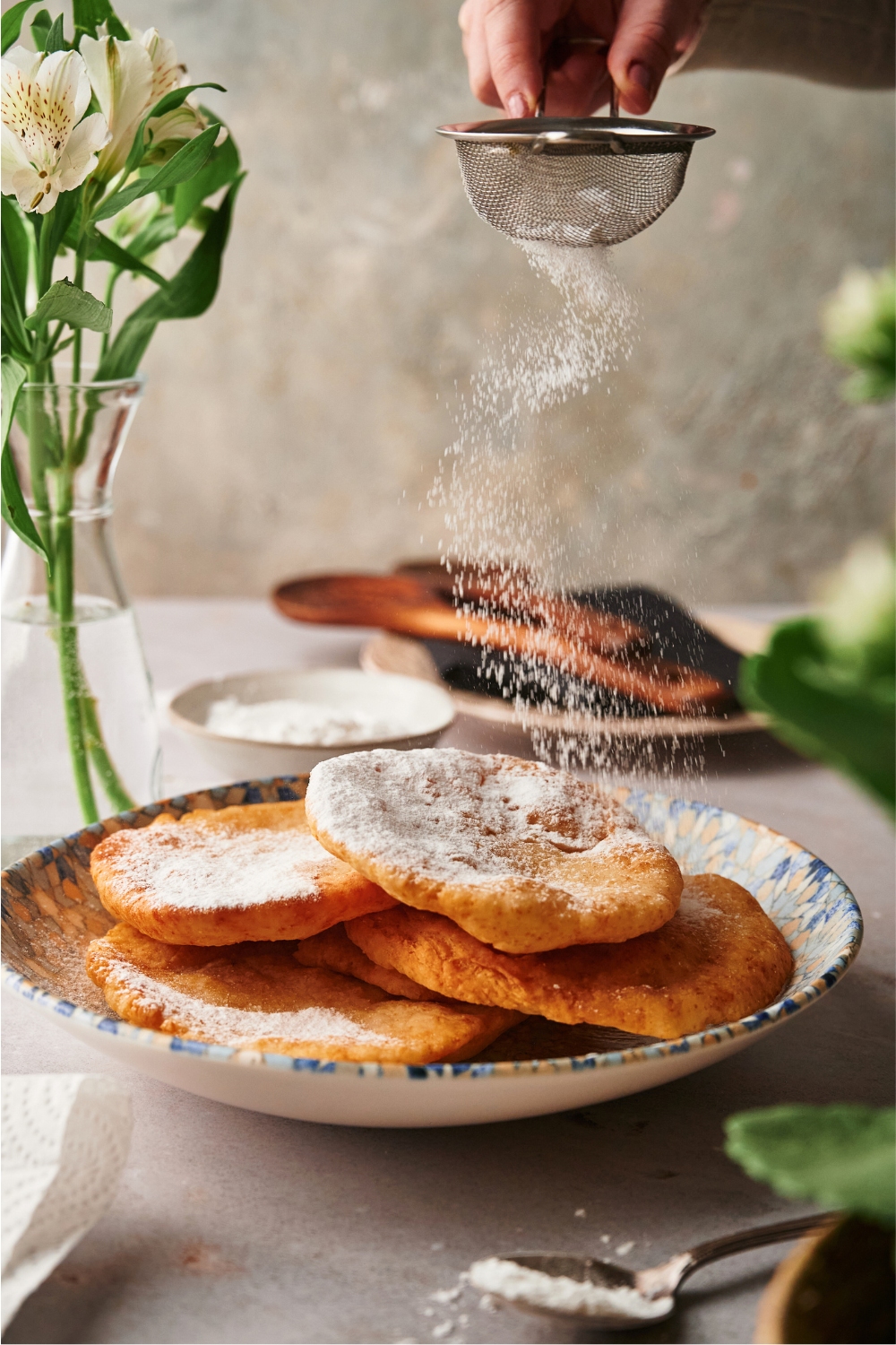 A hand sprinkling powdered sugar on top of a bunch of pieces of fried dough.