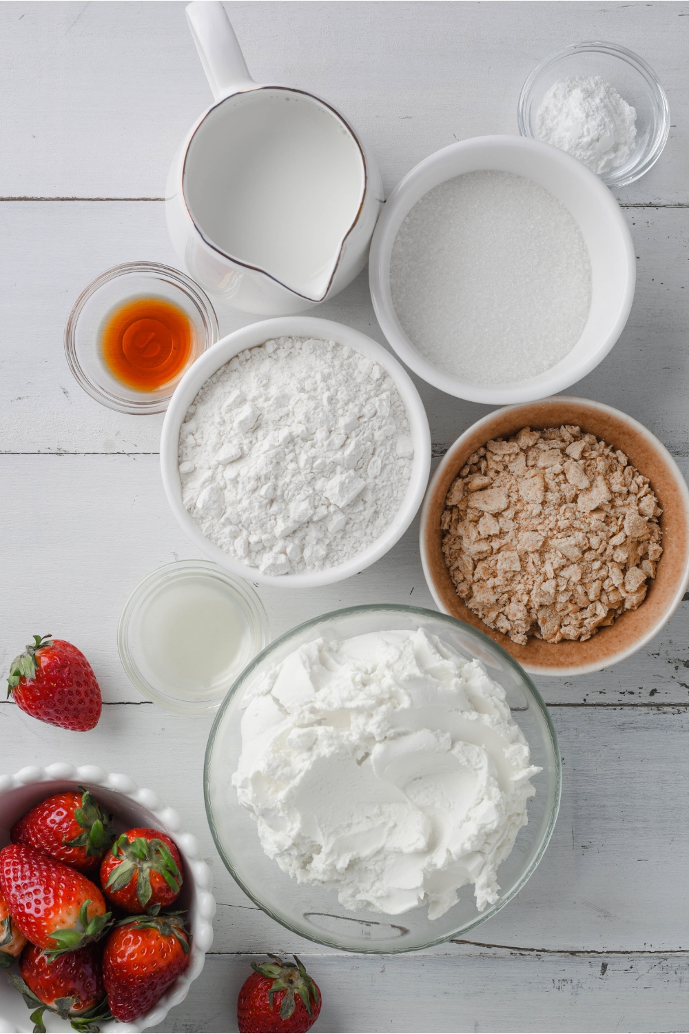 An overhead view of bowls each containing a different ingredient. Such as sugar, flour, Graham cracker crumbs, Cream cheese, Lemon juice, vanilla, baking powder, and milk.