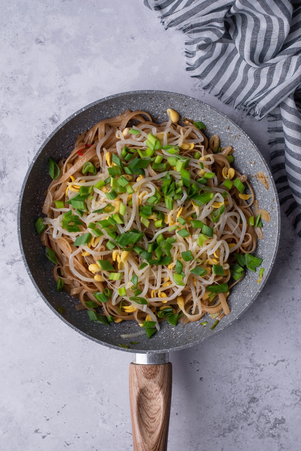 An overhead view of a pan with spicy noodles and chopped green onion and bean sprouts being added to them.