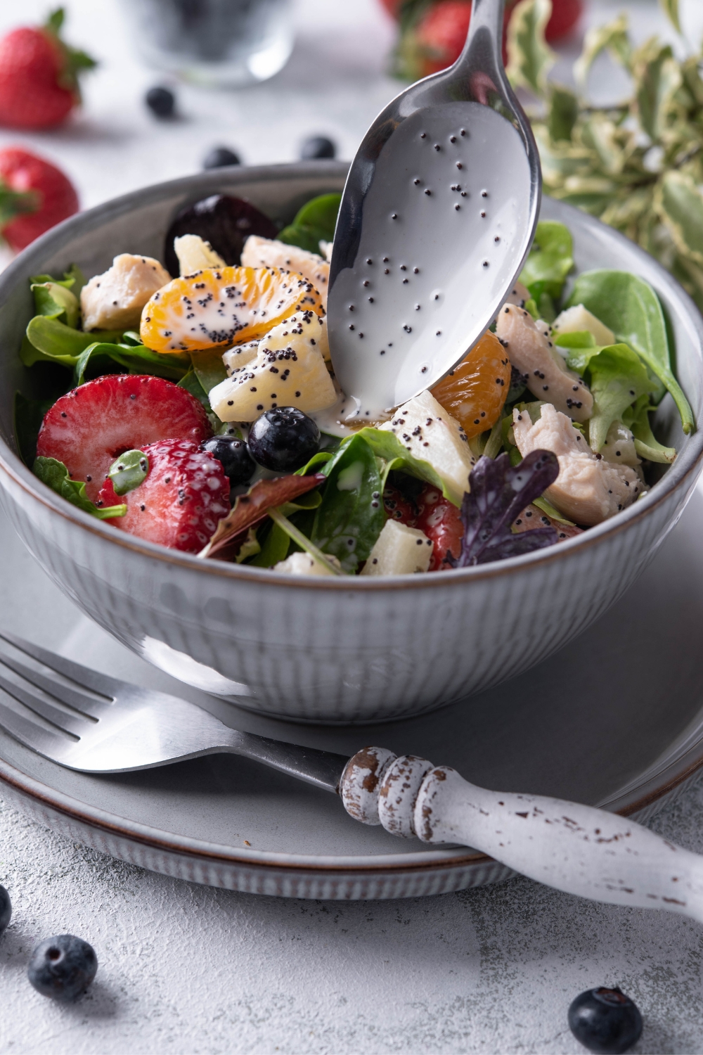 A bowl and saucer serving pair with strawberry poppyseed salad in it. A spoon is drizzling poppyseed dressing on it.
