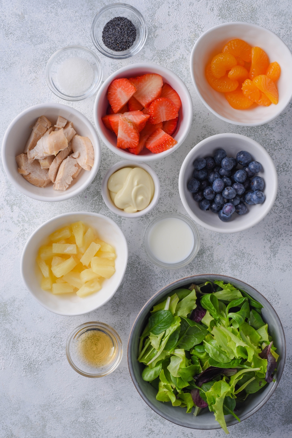 An overhead view of multipole bowls each containing a different ingredient. Pineapple chunks, mixed greens, apple cider vinegar, milk, mayo, blueberries, mandarin oranges, strawberry slices, poppyseeds, sugar, and cooked chicken are all of the ingredients shown.