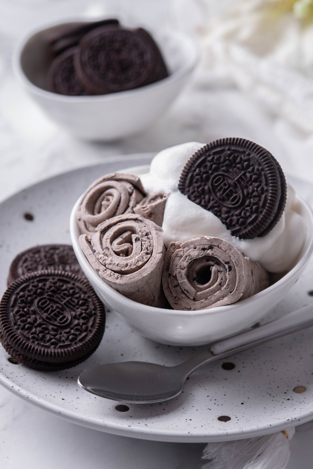 A plate with a bowl with 3 rolled ice creams with whipped cream garnish and a whole oreo.