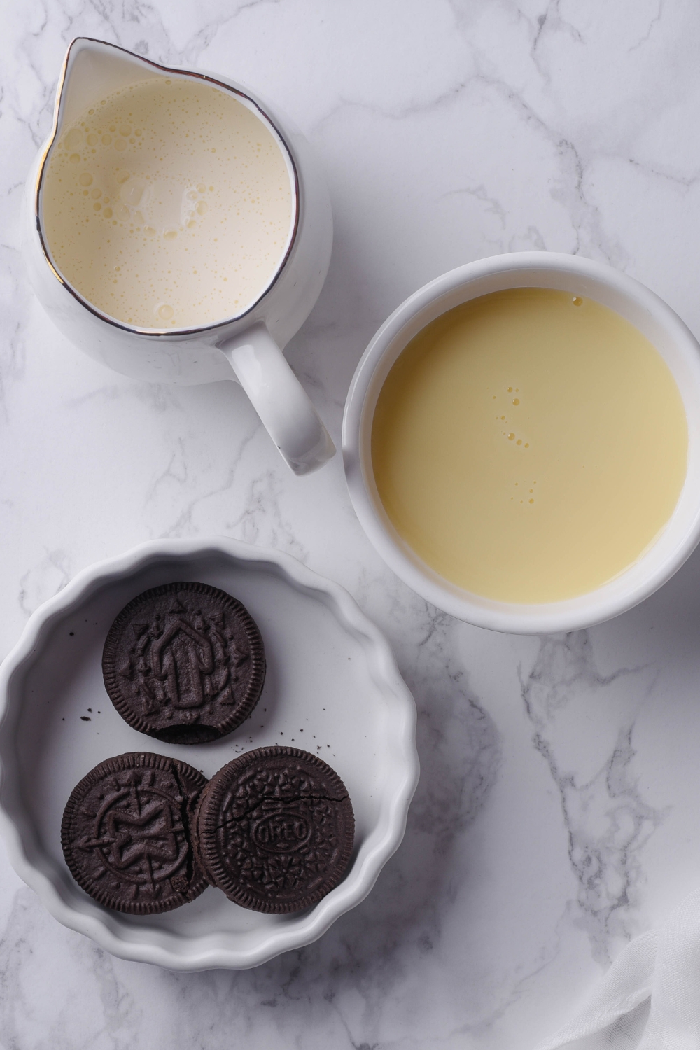 An overhead view of a pitcher with heavy cream, a small bowl with sweetened condensed milk, and a small bowl with 3 oreos in it.
