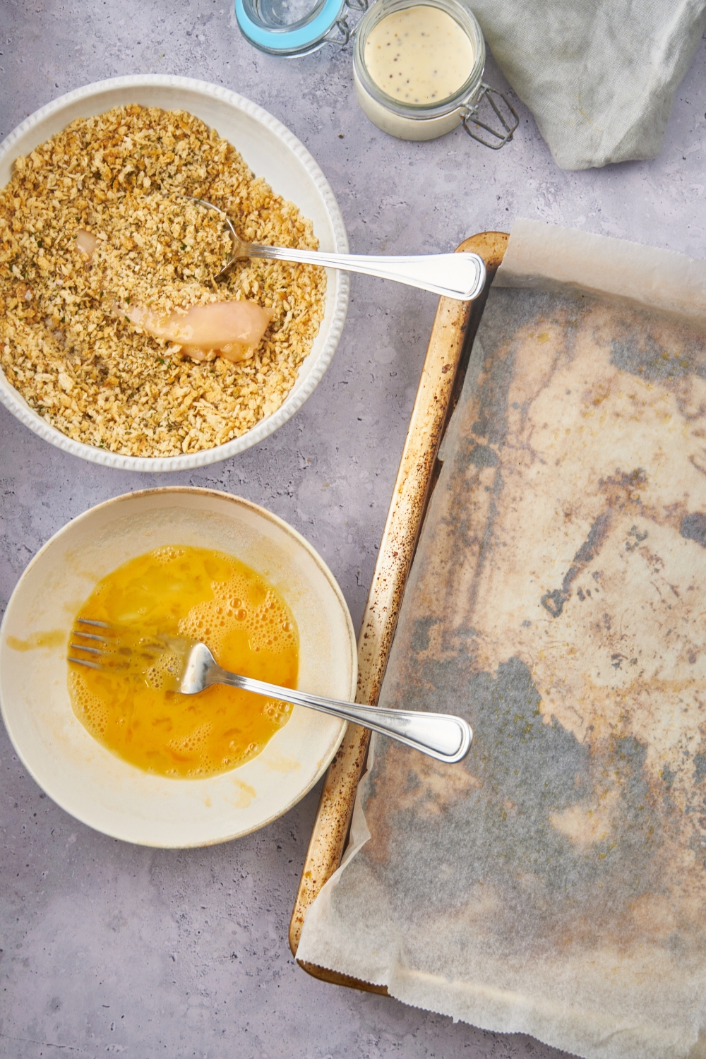 Overhead view of a bowl of panko bread crumbs with a chicken tender being coated using a spoon along with a bowl of beaten eggs and a baking sheet lined with parchment paper.
