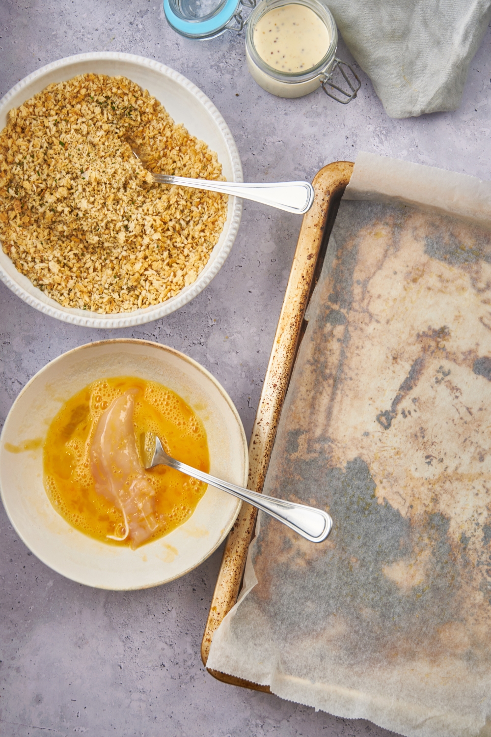 Overhead view of a bowl of panko bread crumbs, a bowl of beaten egg with a chicken tender in it, and a baking sheet lined with parchment paper.