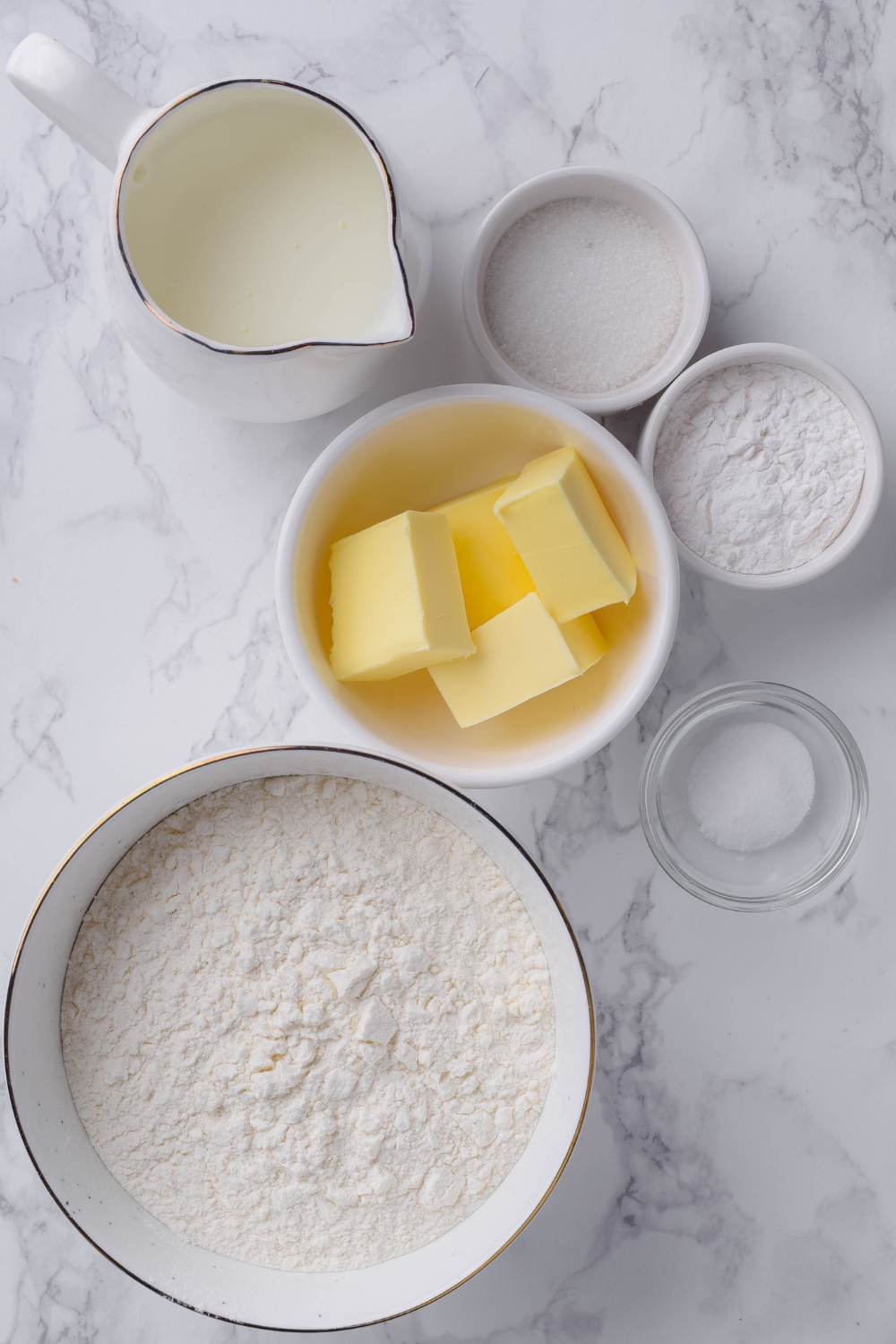 A countertop with a large bowl of flour, cubed butter, salt, sugar, baking soda, and buttermilk.