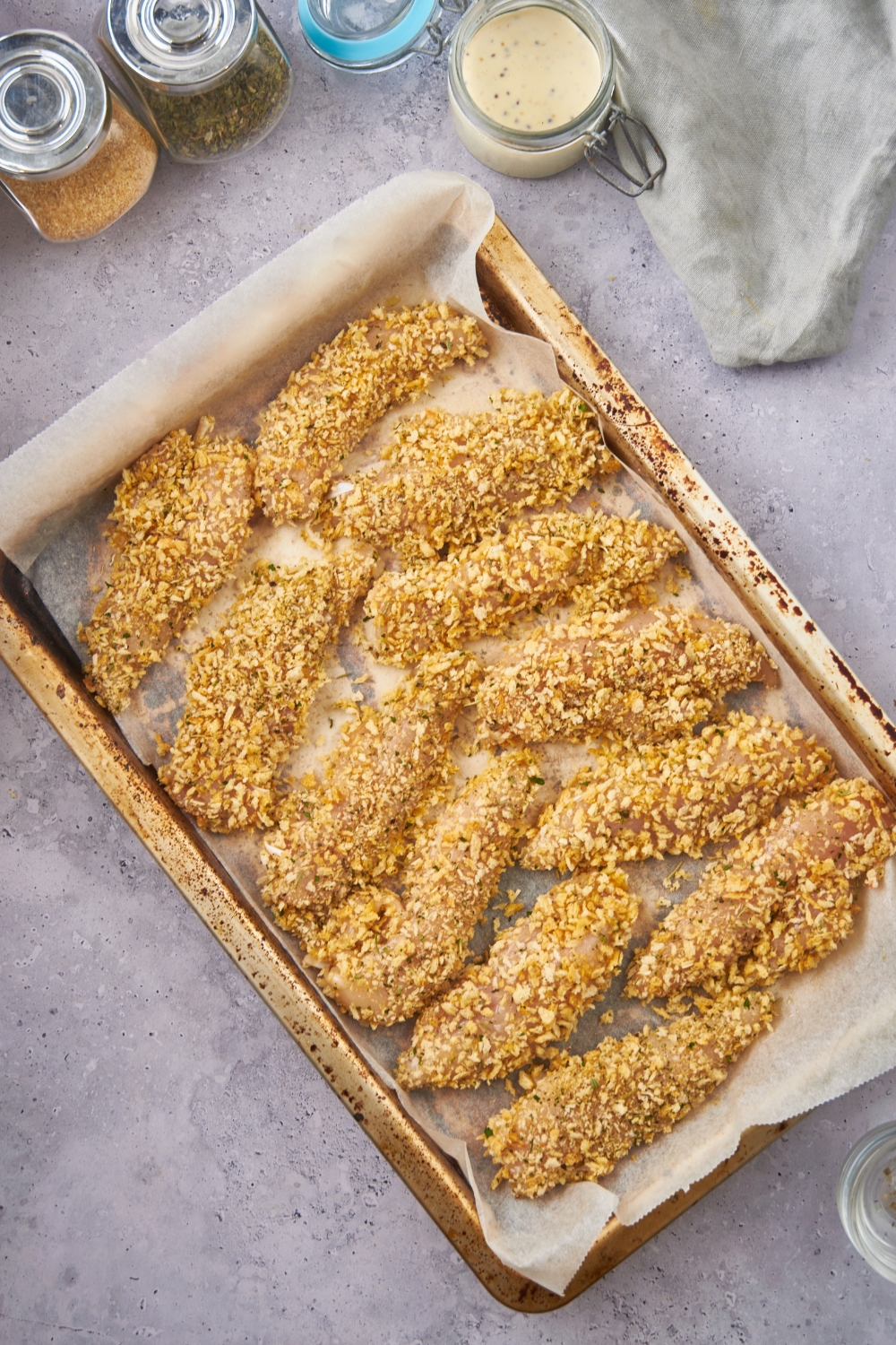 Twelve unbaked chicken tenders coated in panko bread crumbs and lined up on a baking sheet with parchment paper.