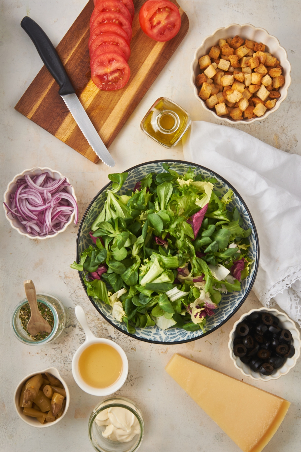 Overhead view of an assortment of ingredients including bowls of lettuce mix, croutons, sliced red onions, black olives, peppers, mayonnaise, a piece of parmesan cheese, sliced tomatoes on a cutting board, and a jar of oil.