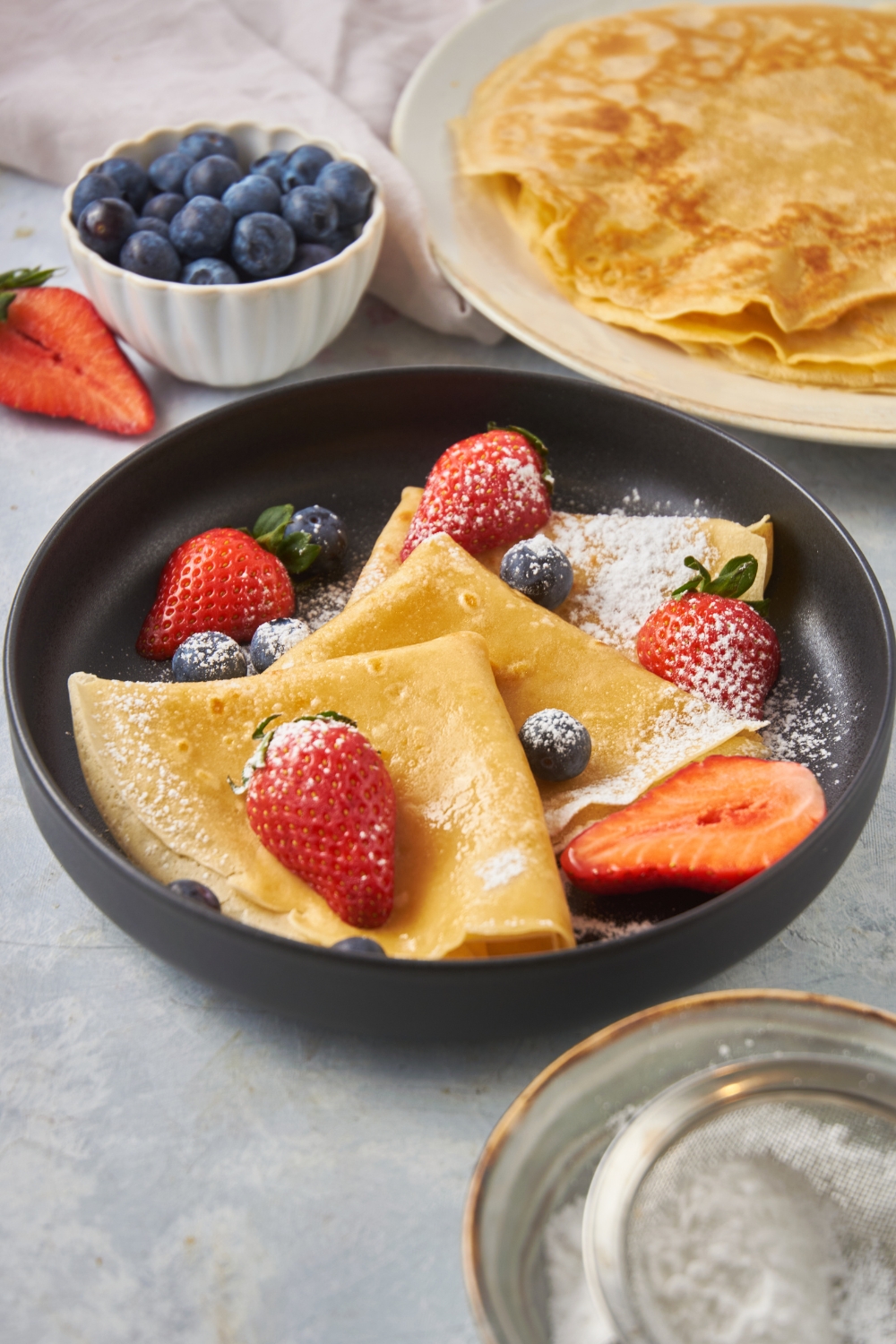 Three crepes layered on top of each other, dusted with powdered sugar and topped with fresh blueberries and halved strawberries.