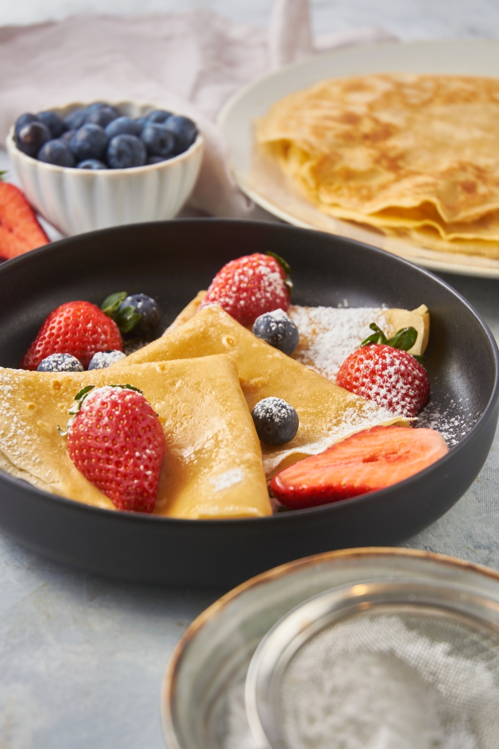 Three crepes layered on top of each other, dusted with powdered sugar and topped with fresh blueberries and halved strawberries.