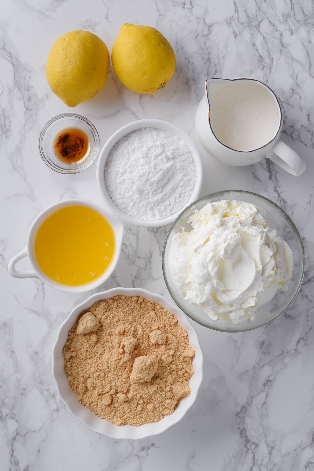 A countertop with a bowl of graham cracker crumbs, melted butter, cream cheese, heavy whipping cream, powdered sugar, vanilla bourbon extract, and two lemons.