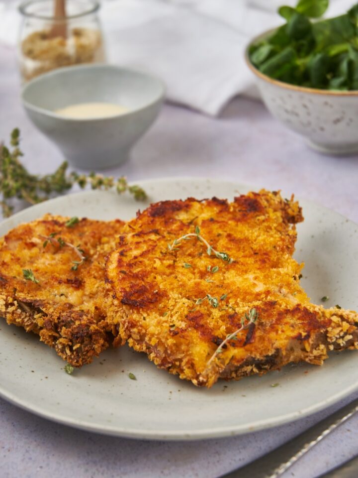 Two golden brown pork chops coated in crispy breading and garnished with fresh green herbs.