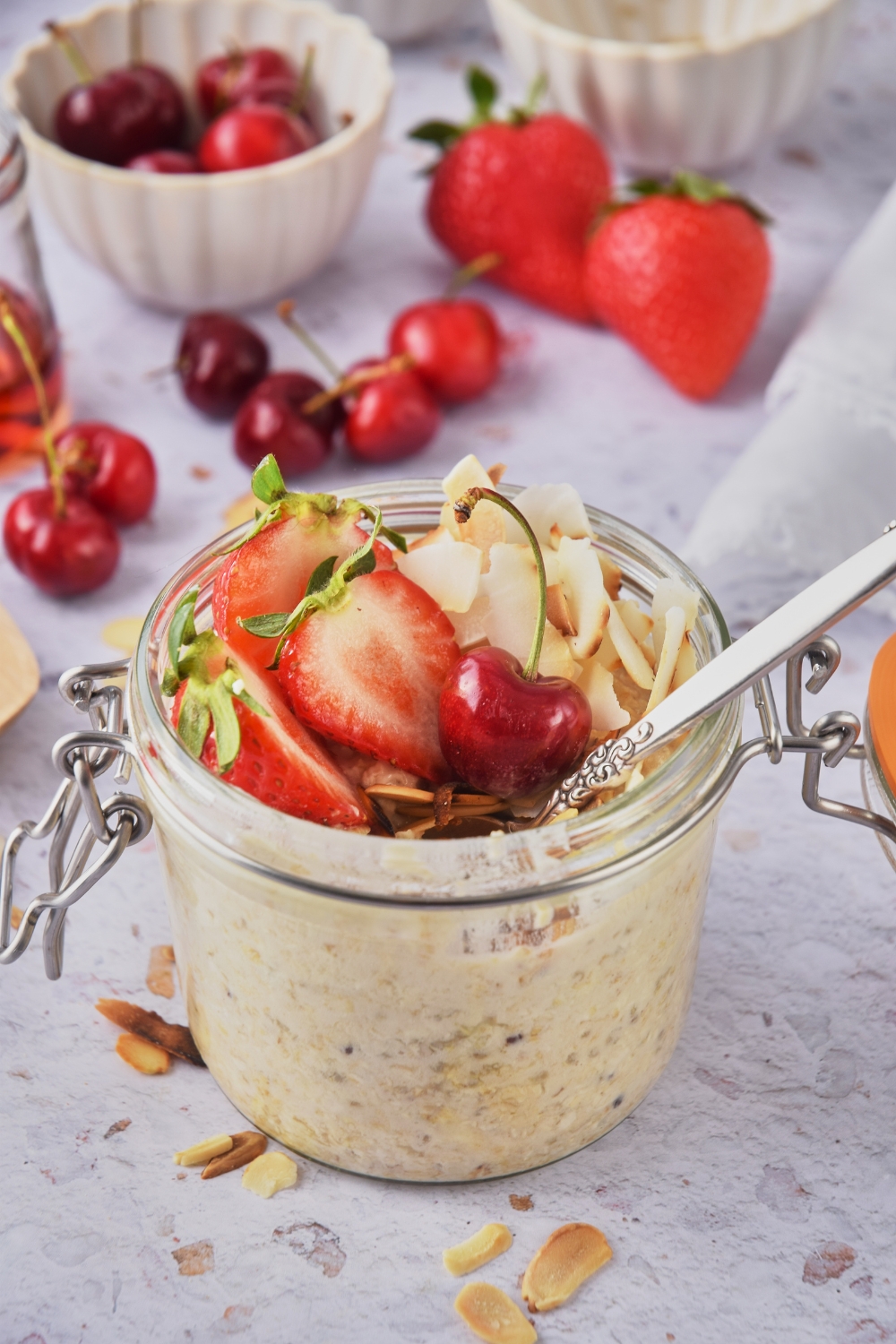 A small mason jar overflowing with overnight oats topped with slices strawberries, cherries, toasted almonds and coconuts. A spoon is in the jar.