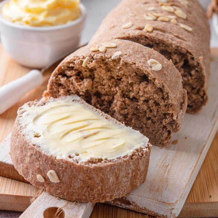 A loaf of brown bread cut into slices with a piece in front of the loaf with butter spread on top.