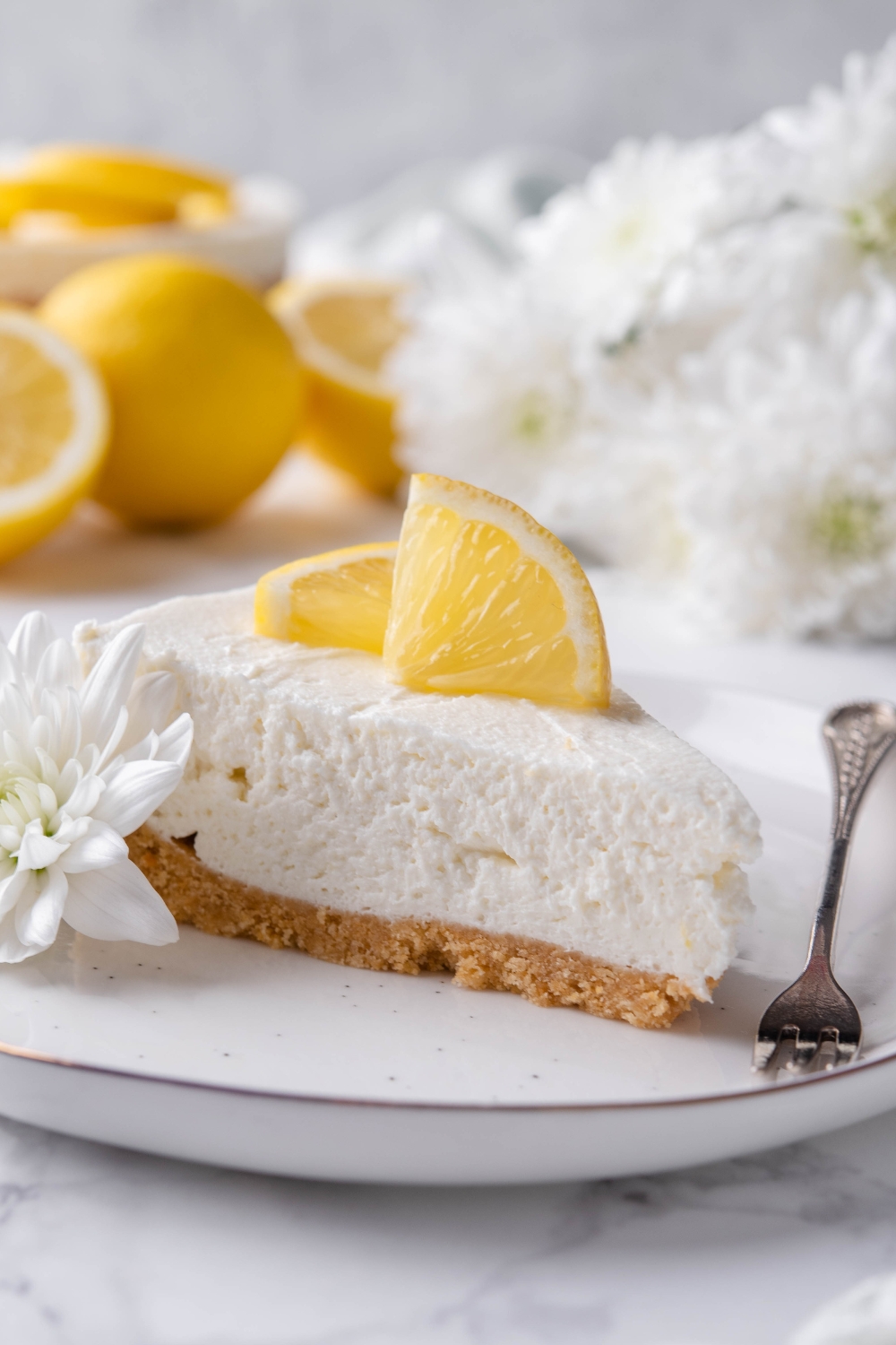A plate with a slice of cheesecake topped with lemon wedges and garnished with a flower. The fork is sitting next to the slice.