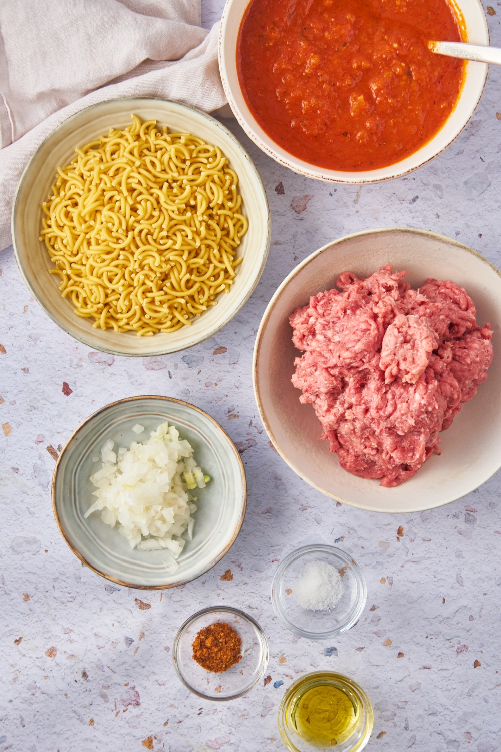 Overhead view of an assortment of ingredients including bowls of raw ground beef, tomato sauce, dried macaroni noodles, diced onion, oil, salt, and spices.