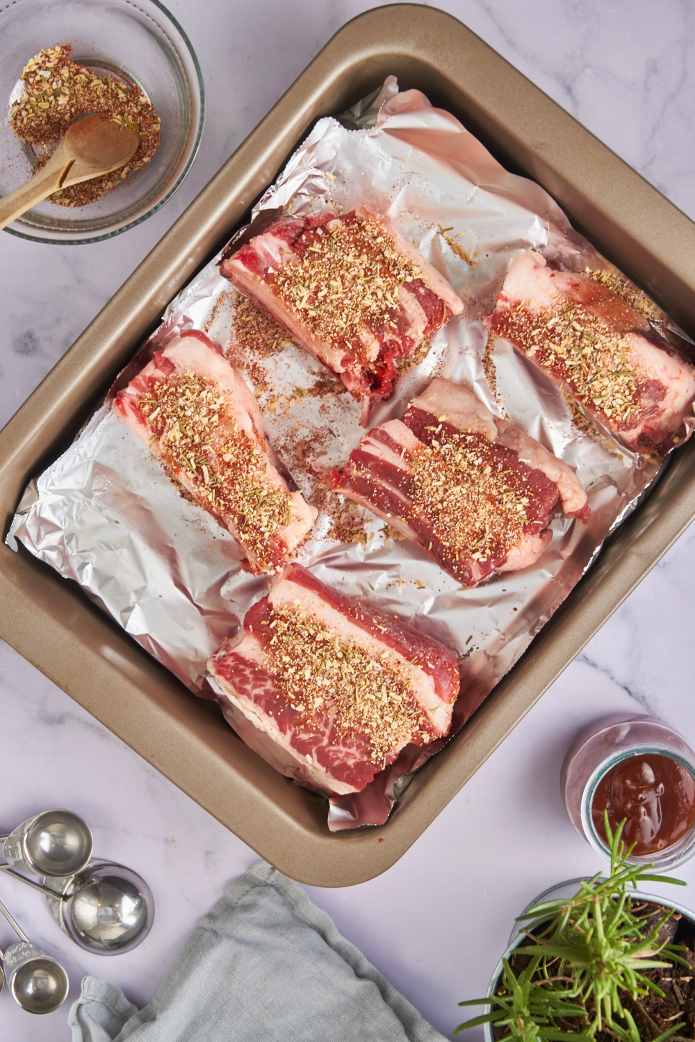 Five beef back ribs covered in a spice blend spread evenly on a baking sheet lined with foil.