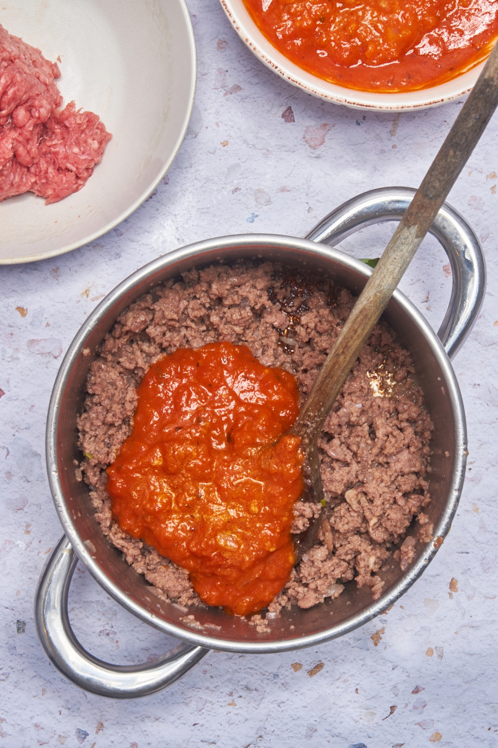 A large silver pot with two handles filled with cooked ground beef, chunky tomato sauce, and a wooden spoon.