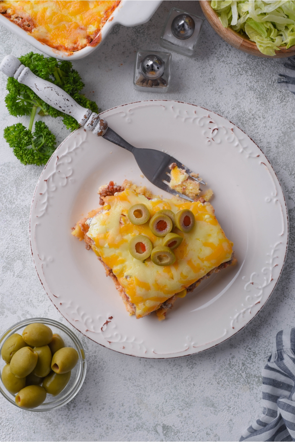 Overhead view of a serving of Mexican cornbread casserole with a garnish of sliced green olives and a fork on the plate. A bite is missing from the casserole.