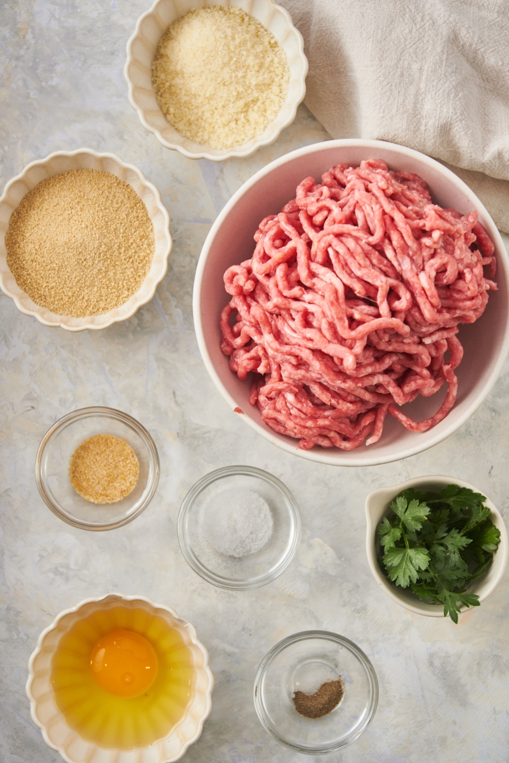 Overhead view of an assortment of ingredients including bowls of raw ground beef, an egg, green herbs, spices, bread crumbs, and parmesan cheese.