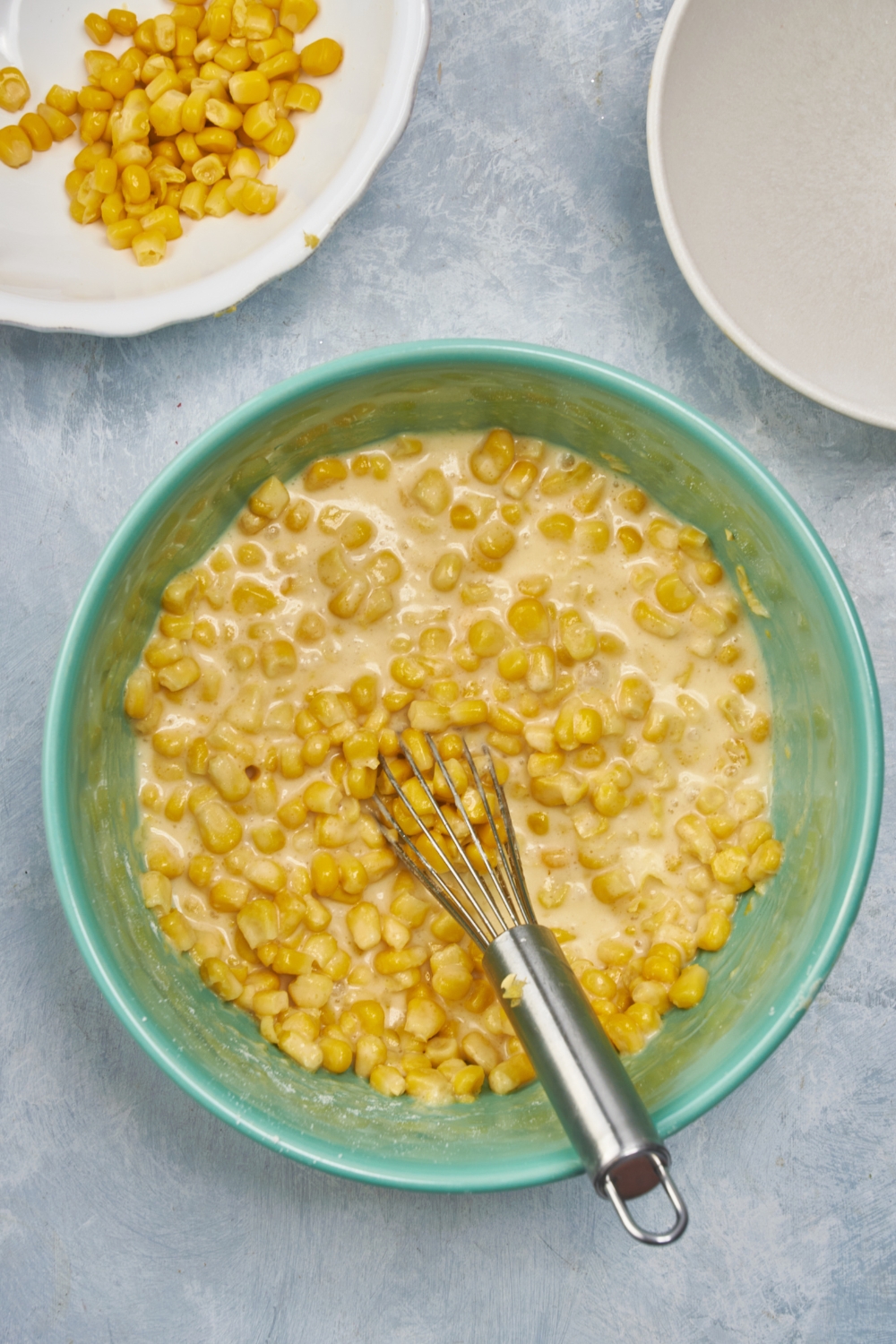 A blue bowl filled with wet ingredients and whole corn kernels added but not fully mixed together. There is a whisk in the bowl.