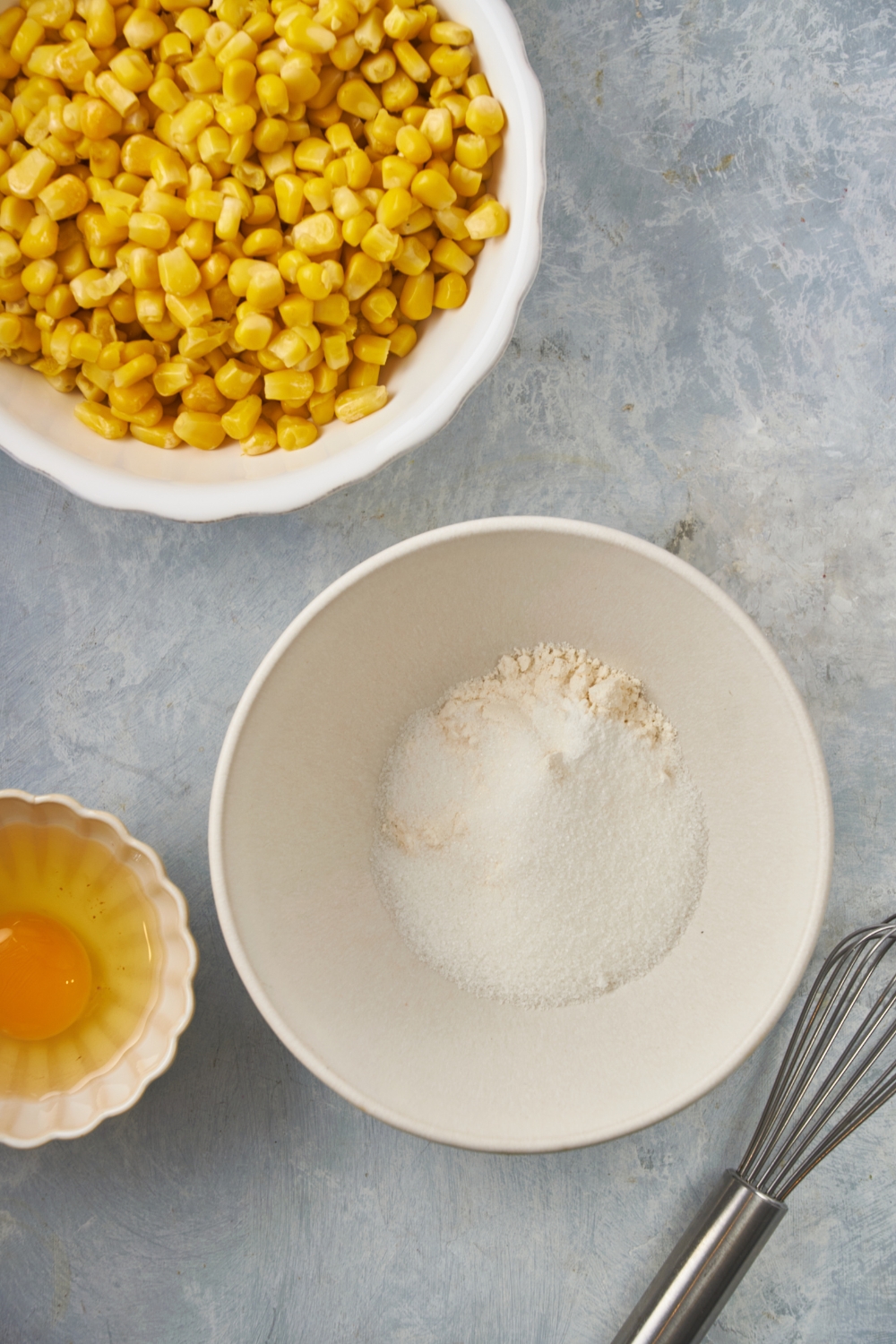 A white bowl with flour and sugar added but not mixed together. Surrounding the bowl is a whisk, an unbeaten egg, and a bowl of corn.