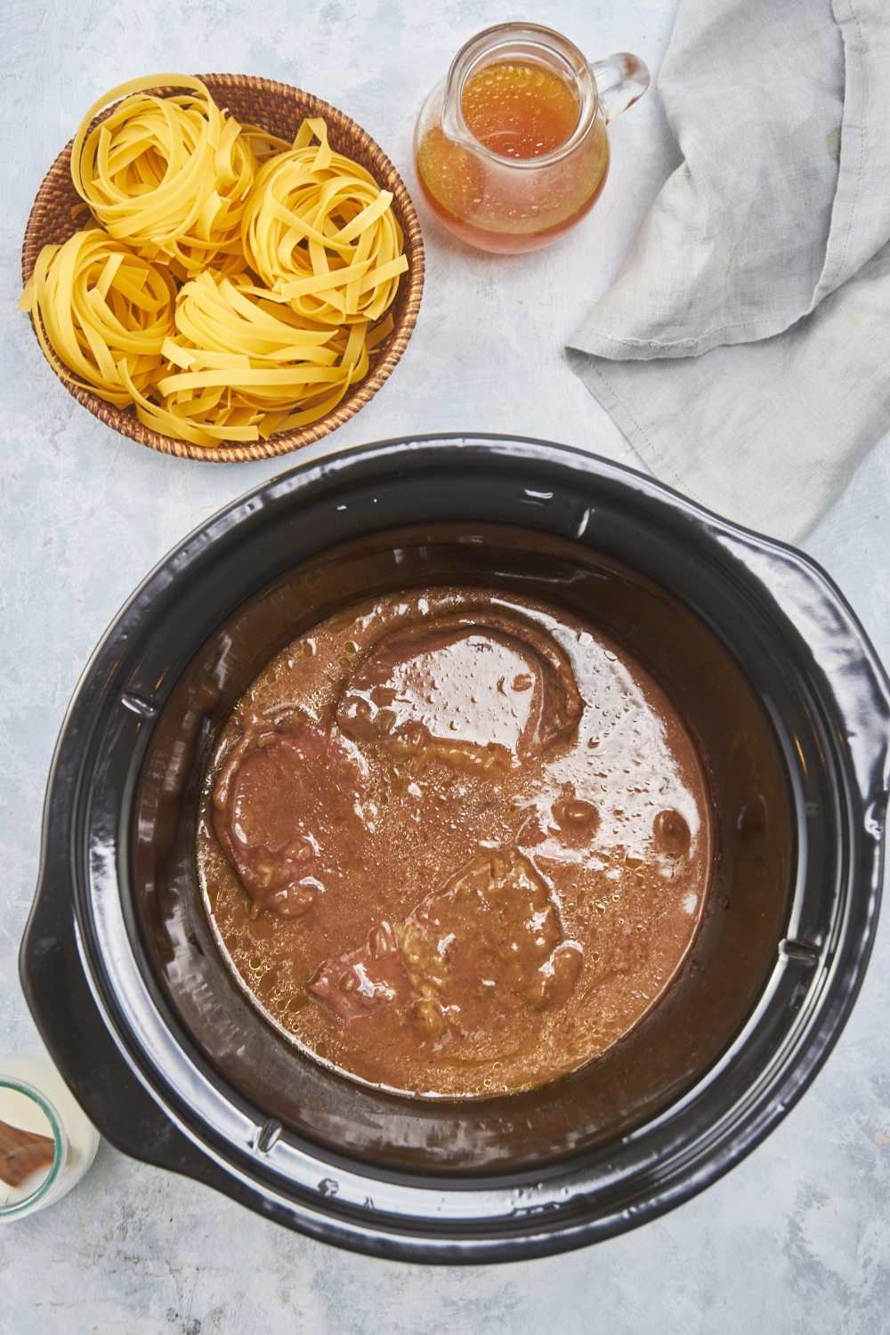 Black crockpot with three cooked steaks covered in a brown sauce. The crockpot is next to a bowl of dried pasta and a jar of broth.