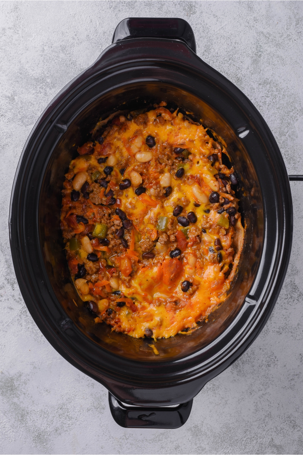 A black slow cooker filled with cooked ground beef, beans, crushed tomatoes, peppers, onions, and melted cheese.