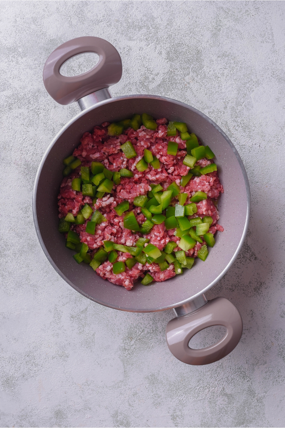 A grey sauce pot filled with raw ground beef and diced green peppers.