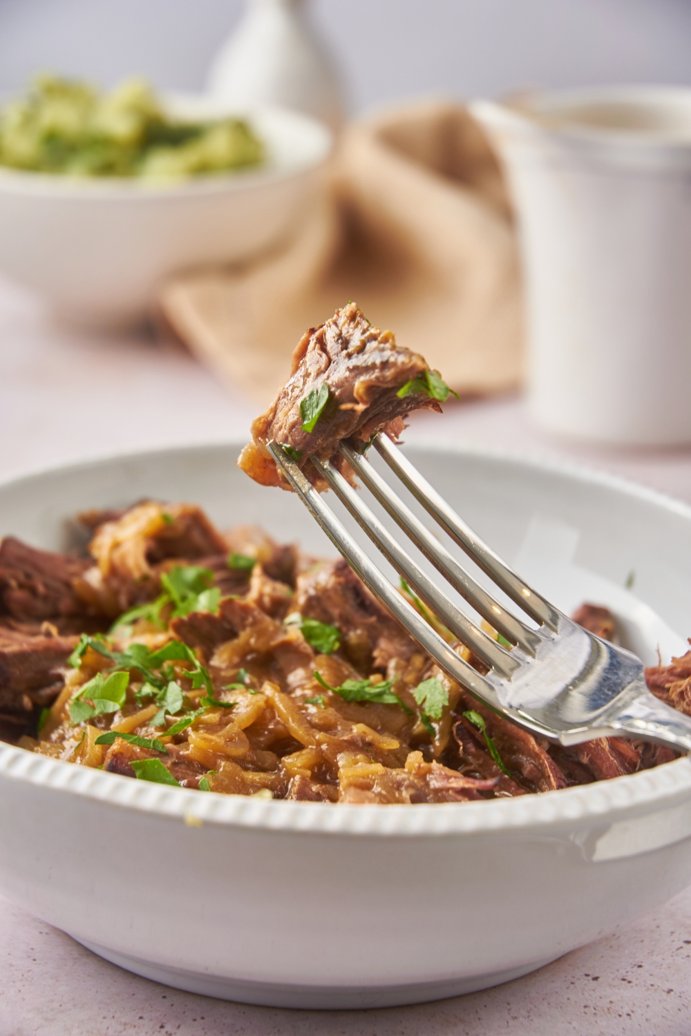 A fork holding a piece of beef roast with bits of green herbs garnished on top. In the background is a bowl of shredded beef.