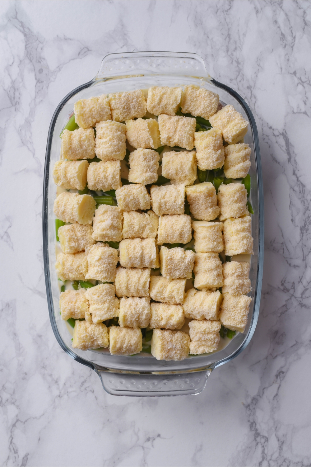 A clear baking dish layered with green beans and frozen tater tots on top.