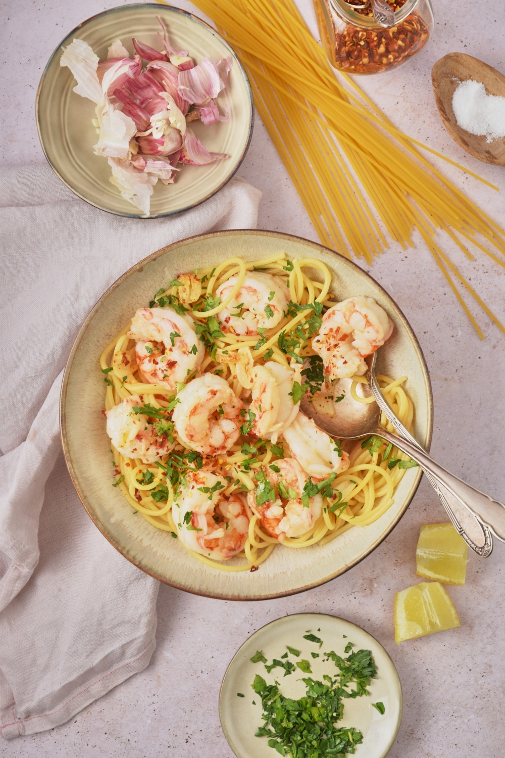 Overhead view of a bowl of shrimp spaghetti garnished with fresh green herbs. There is a spoon and fork in the bowl and the bowl is surrounded by raw ingredients.