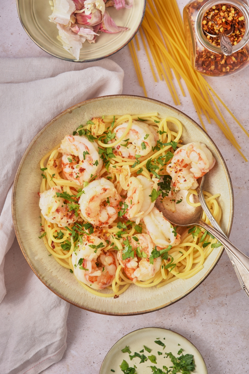 Overhead view of a bowl of shrimp spaghetti garnished with fresh green herbs. There is a spoon and fork in the bowl and the bowl is surrounded by raw ingredients.