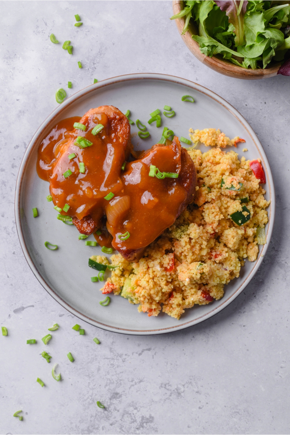 Overhead view of two pork chops covered in a brown gravy with a garnish of green onions and a side of couscous salad.