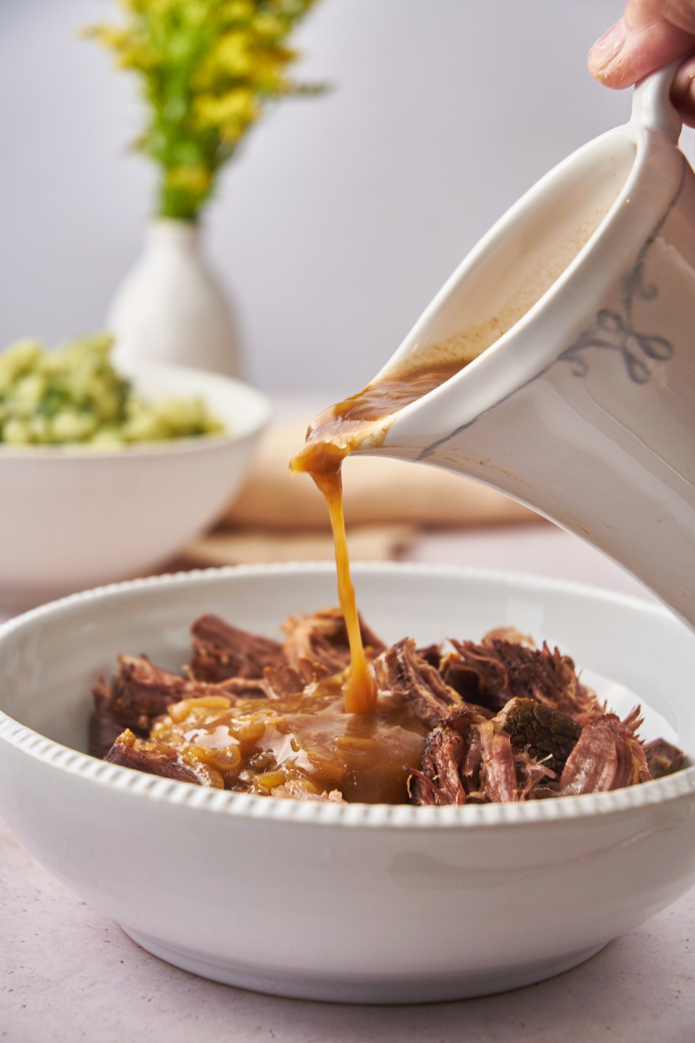 A bowl of cooked and shredded eye of round roast with a pitcher of au jus being poured over top of the meat.