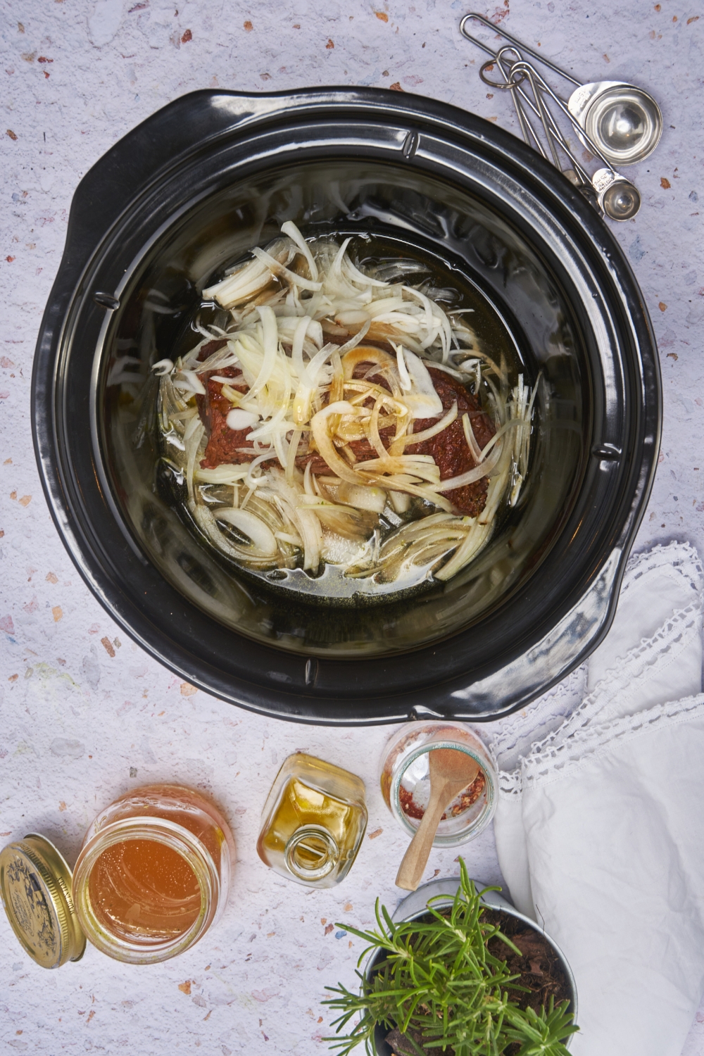 A black crockpot layered with thinly sliced onions, seasoned steak, more onions piled on top, and a brown sauce poured over top of the onions.