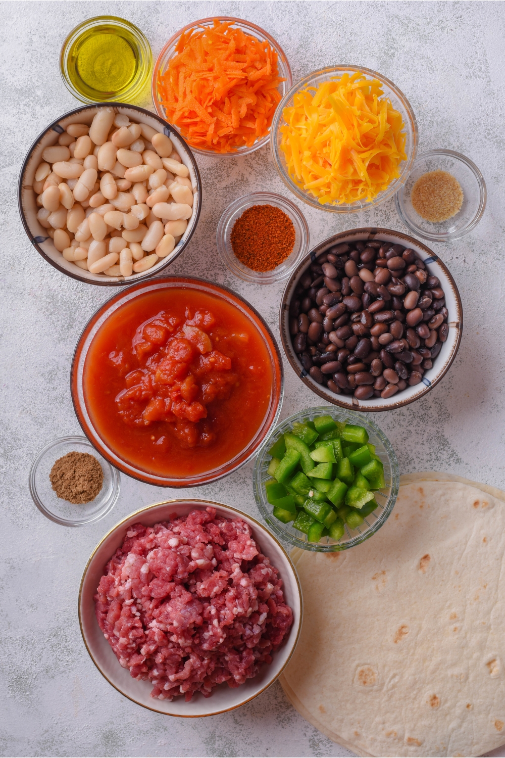 An assortment of ingredients including bowls of crushed tomatoes, black beans, white beans, grated carrots, shredded cheese, oil, spices, ground beef, and a stack of tortillas.