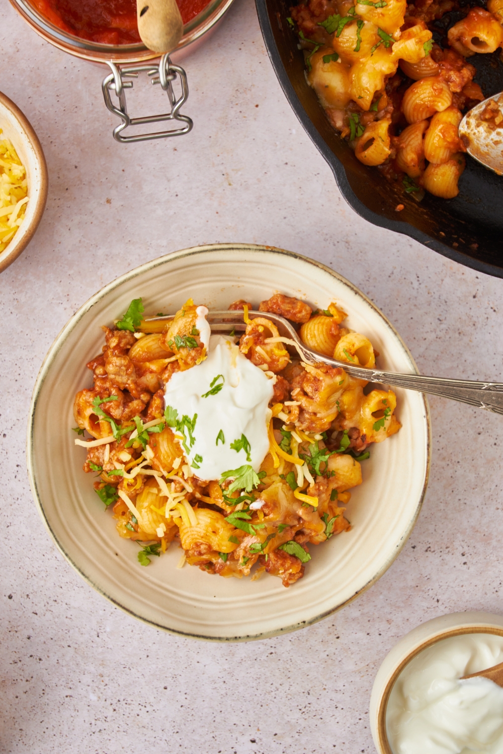 A bowl of taco pasta casserole topped with shredded cheese, fresh green herbs, and sour cream.