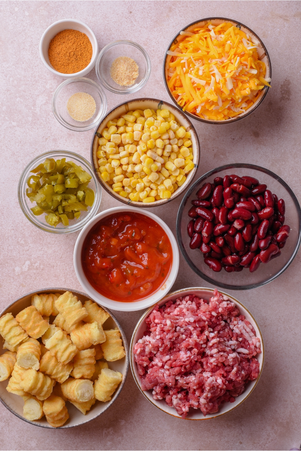 An assortment of ingredients including bowls of shredded cheese, corn, black beans, enchilada sauce, raw ground beef, tater tots, jalapeños, and spices.