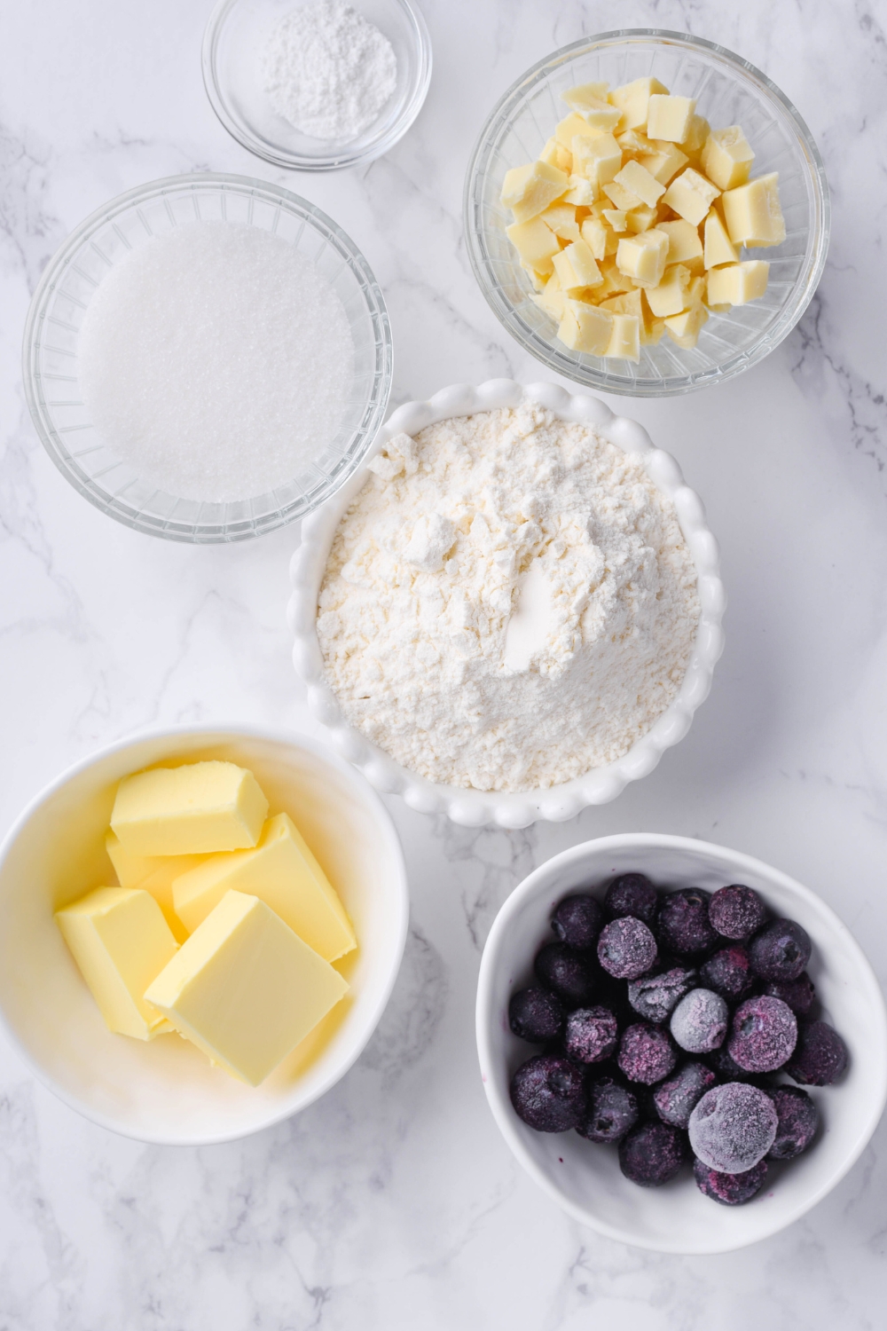 A countertop with flour, butter, blueberries, white chocolate chunks, sugar, and baking powder in separate bowls.
