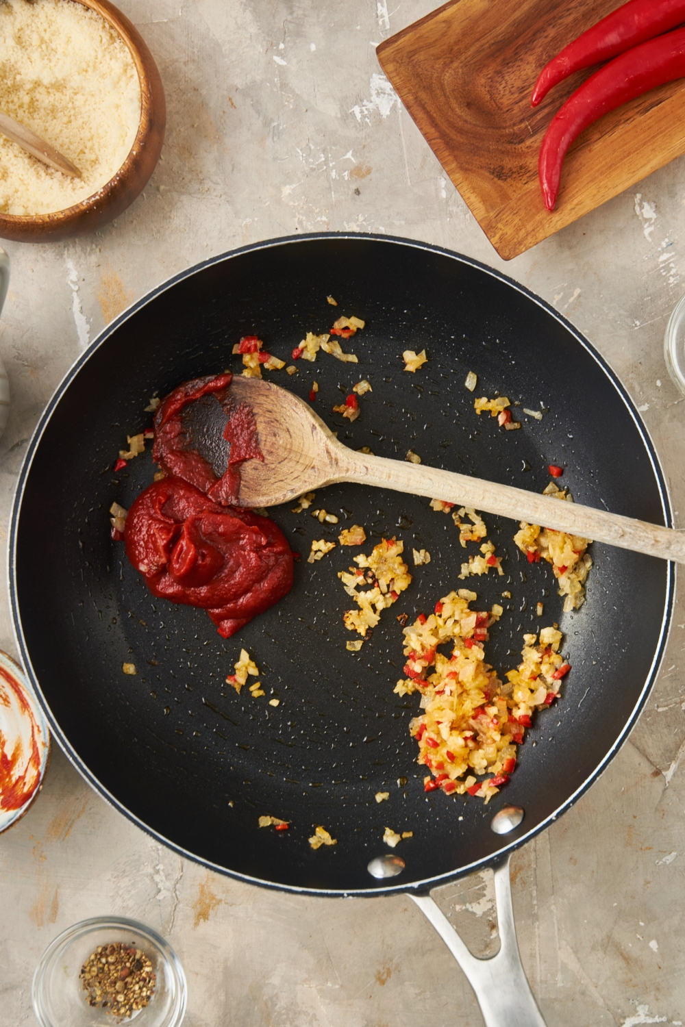 Black skillet with minced garlic, diced chilies, and a dollop of tomato paste added using a wooden spoon.