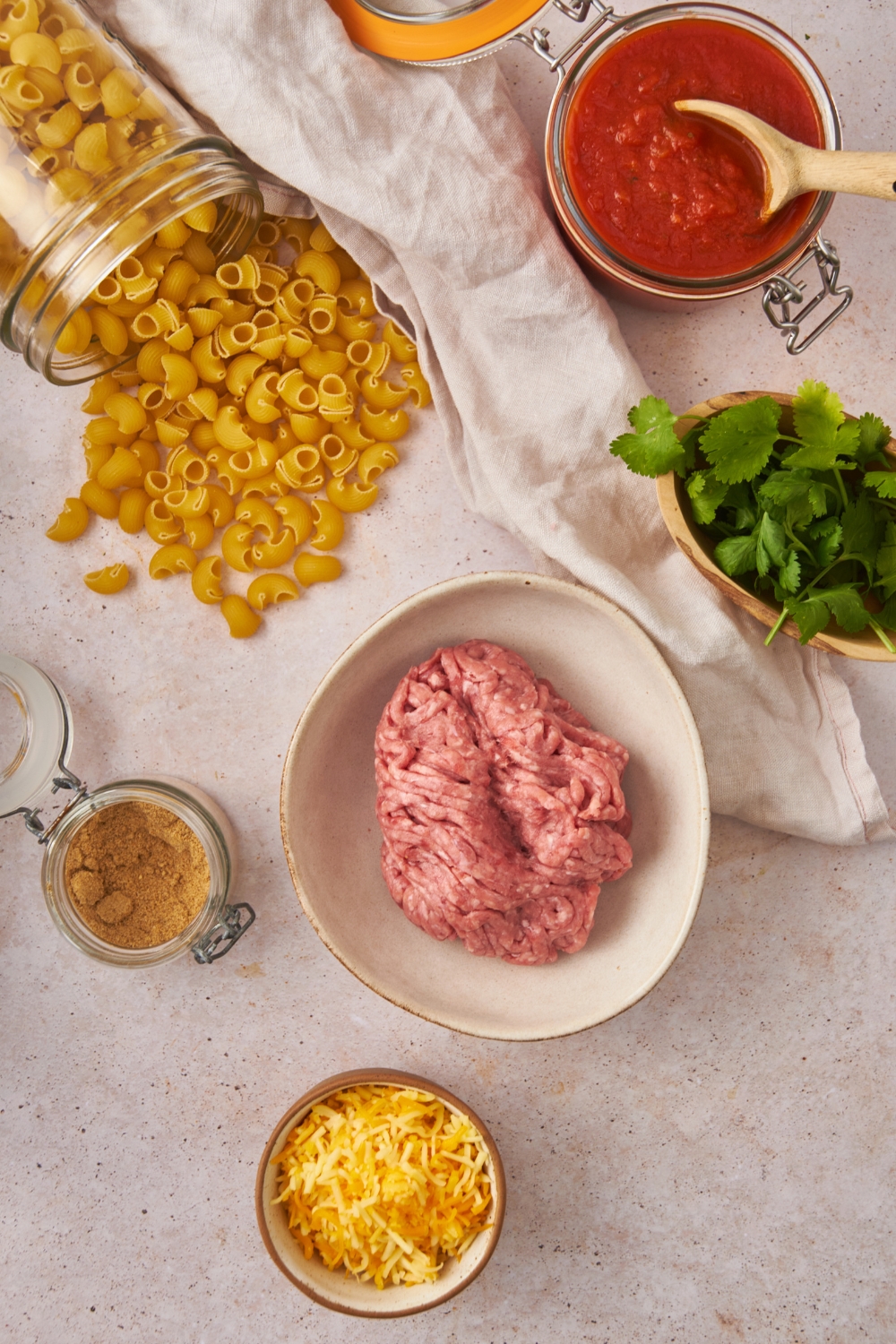 Overhead view of an assortment of ingredients including bowls of raw ground beef, salsa, fresh herbs, shredded cheese, taco seasoning, and a container of dried pasta spilling onto the counter.