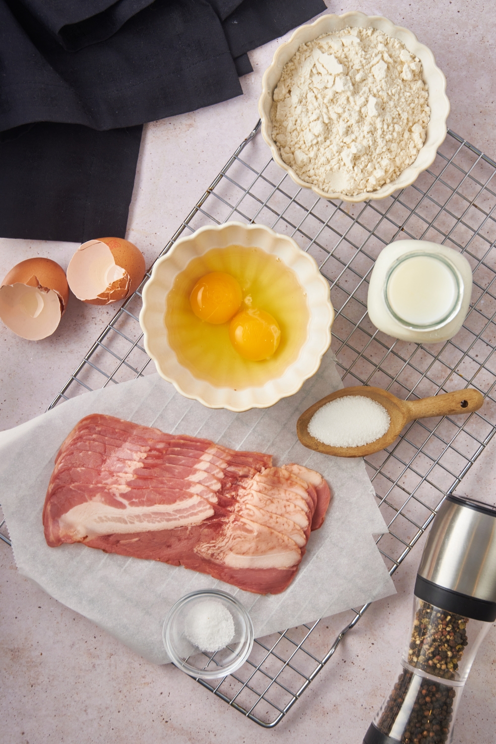 Overhead view of an assortment of ingredients including bowls of eggs, milk, flour, a pepper shaker, a spoonful of sugar, and a package of raw bacon over top of a piece of wax paper.