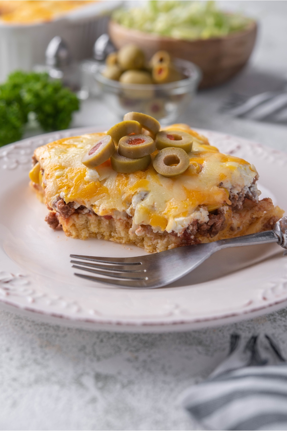 A serving of cornbread taco casserole on a white plate with a garnish if sliced green olives and a fork is on the plate.