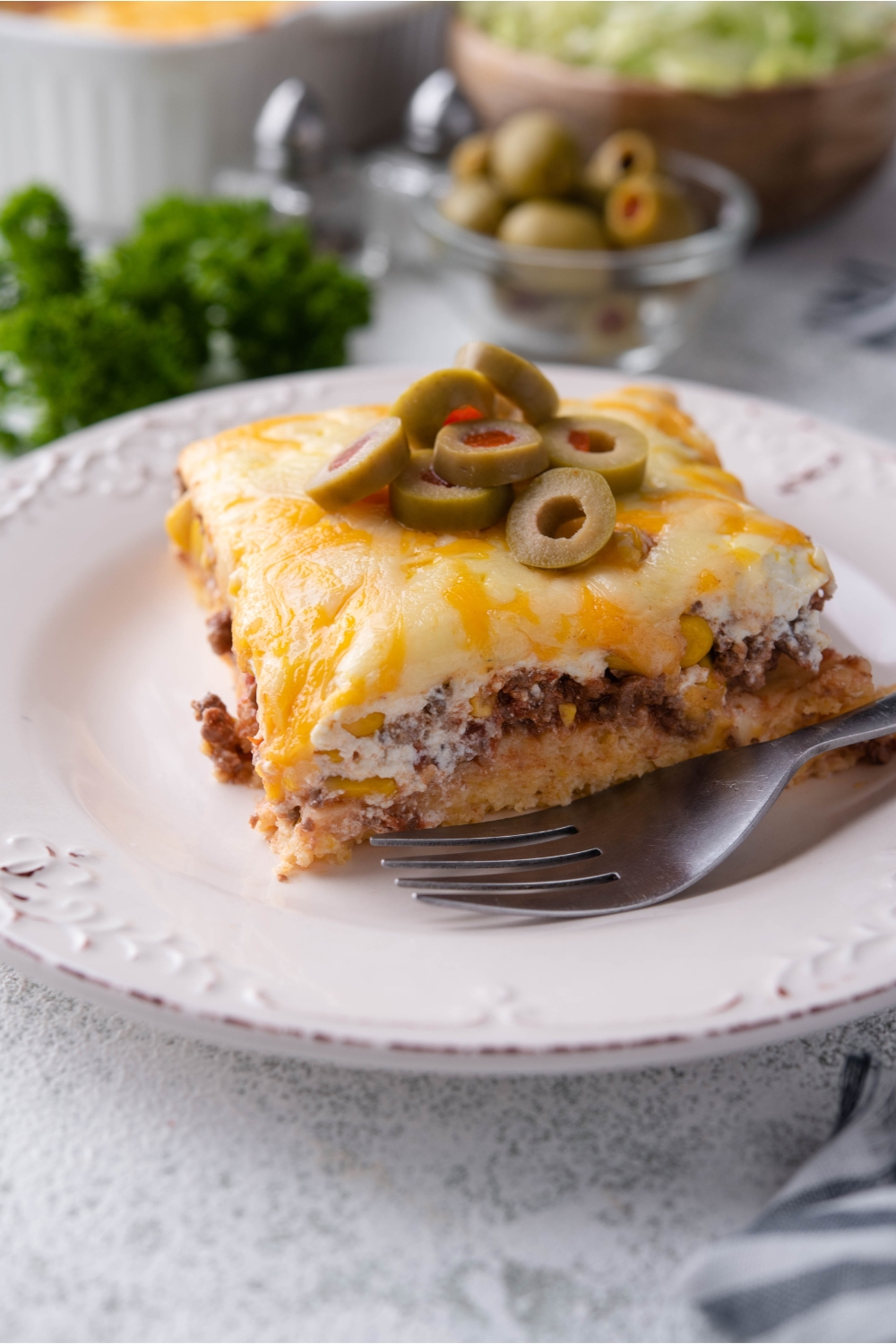 A serving of cornbread taco casserole on a white plate with a garnish if sliced green olives and a fork is on the plate.