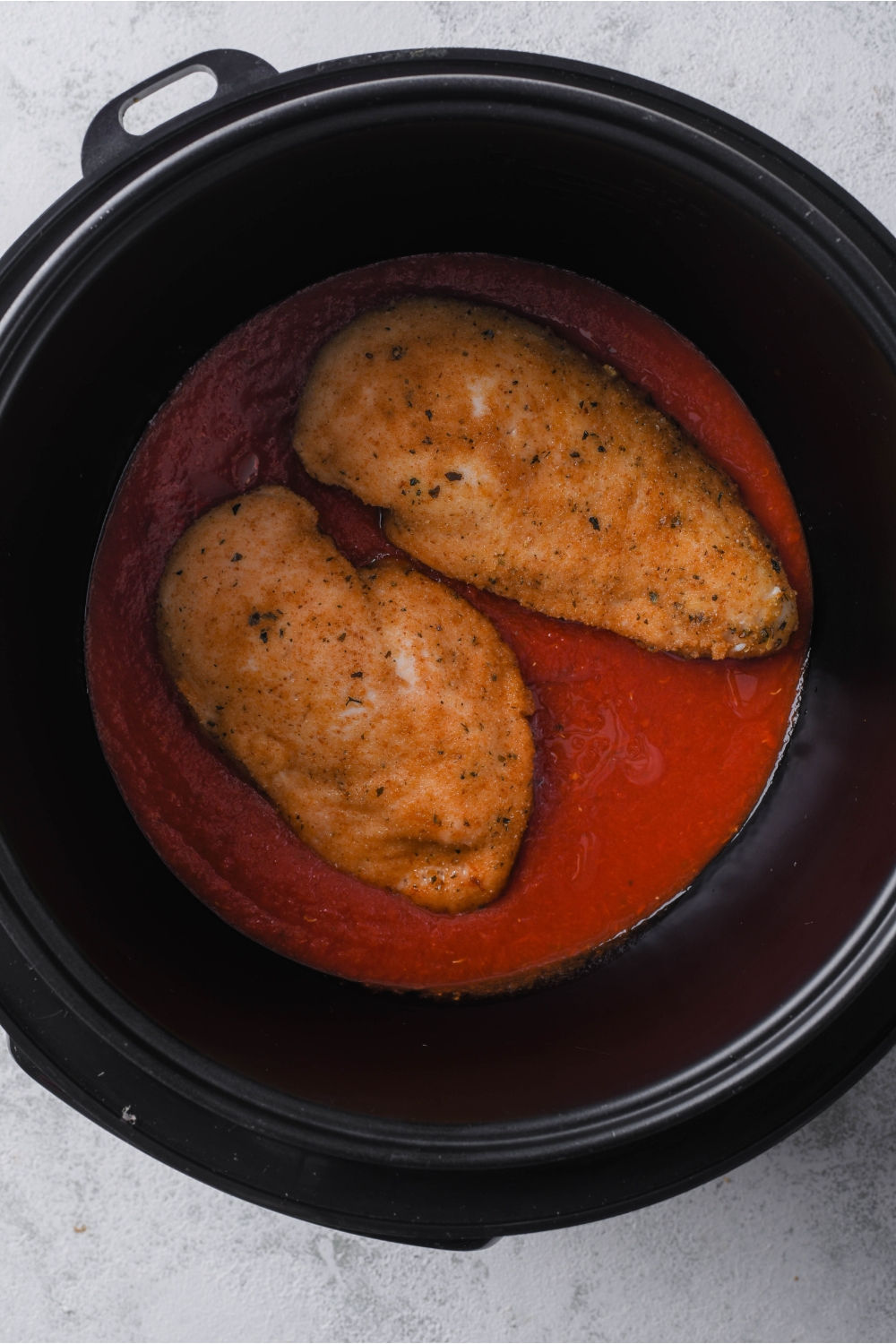 Two seasoned and golden brown chicken breasts in a black crockpot on top of marinara sauce.