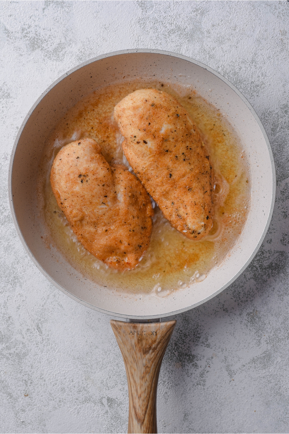 Two seasoned chicken breasts frying in oil in a grey skillet.