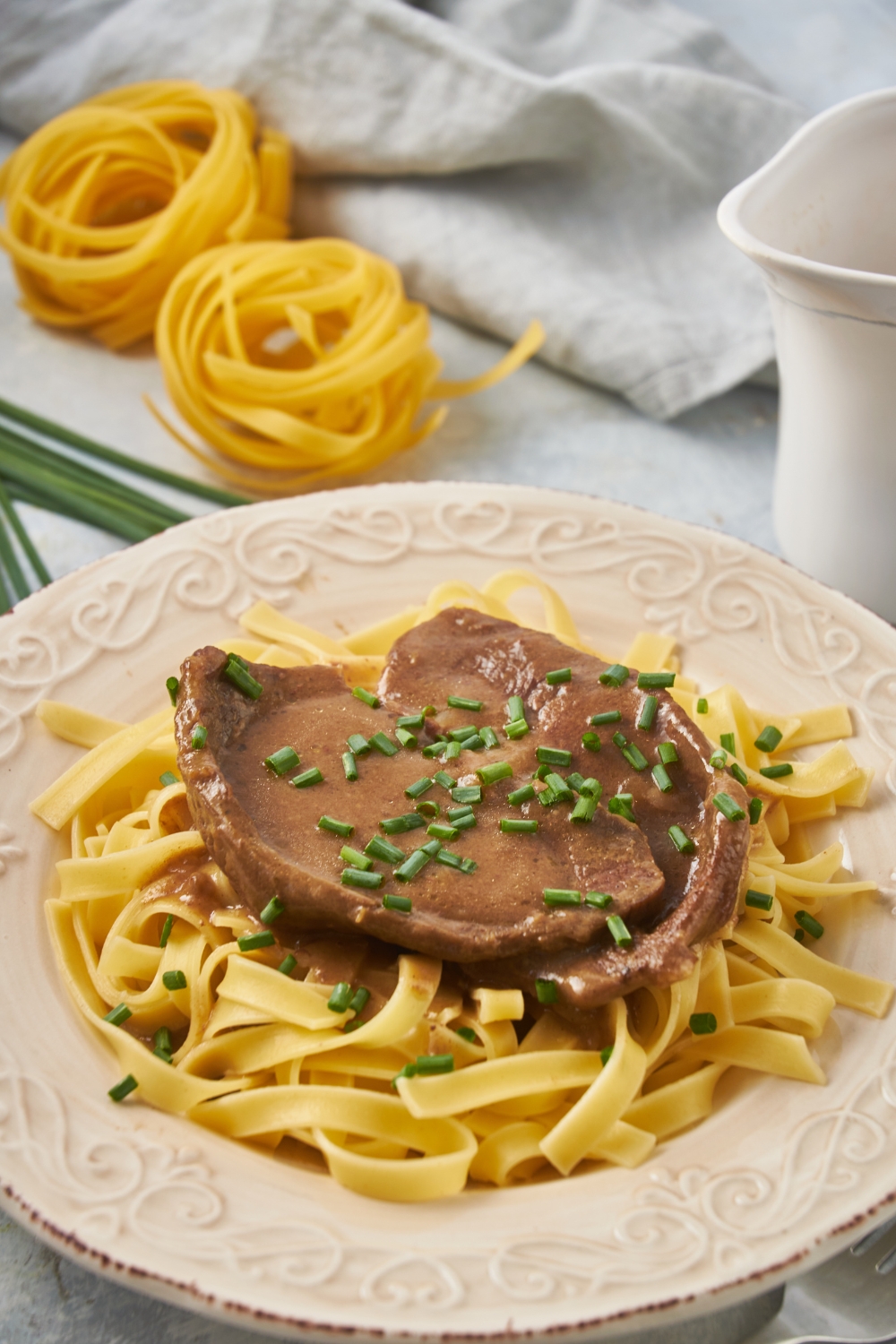 A cube steak covered in brown gravy garnished with sliced green onion and served on a bed of linguini noodles.