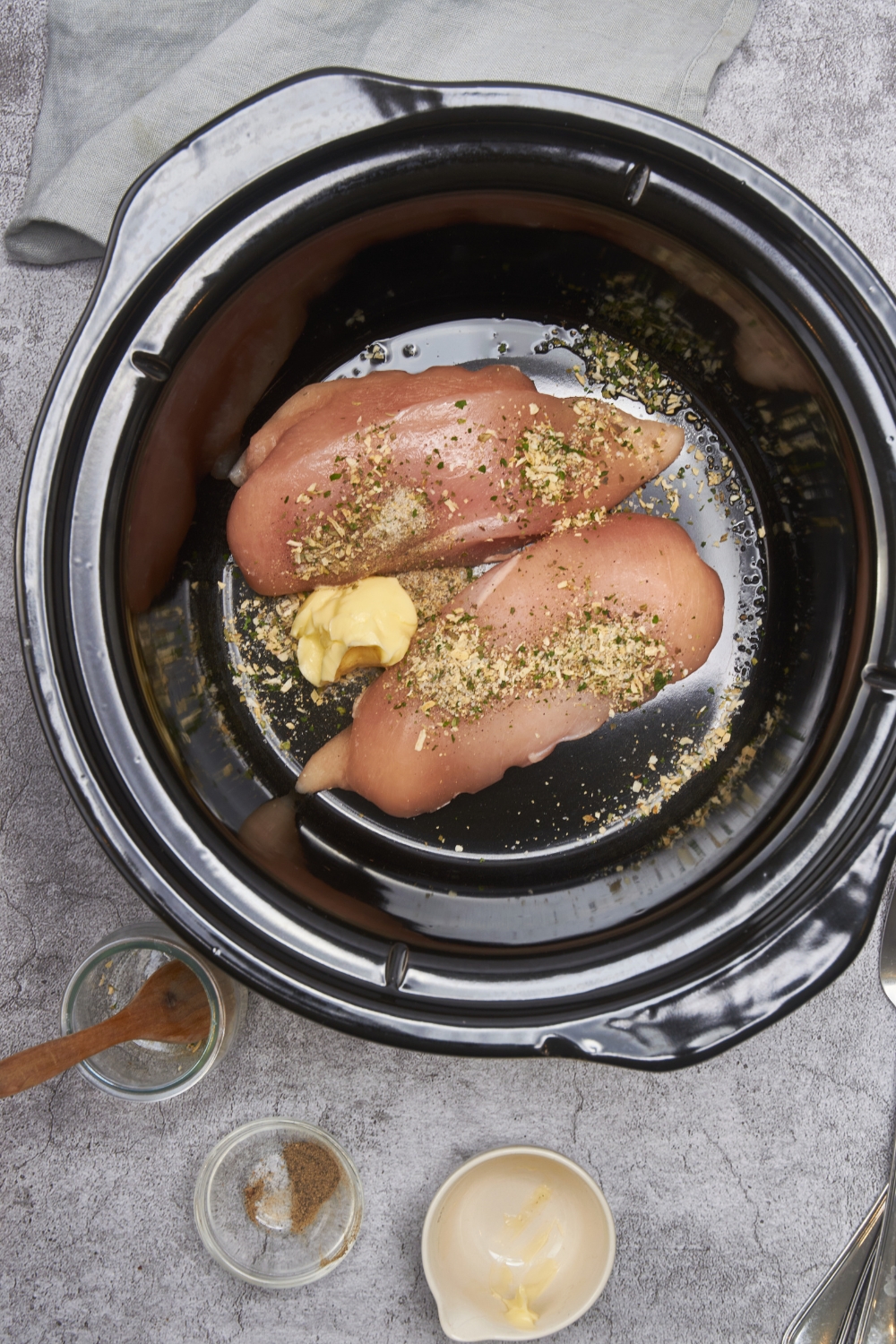 Two raw chicken breasts covered in seasoning in the bottom of a crockpot with a scoop of butter.